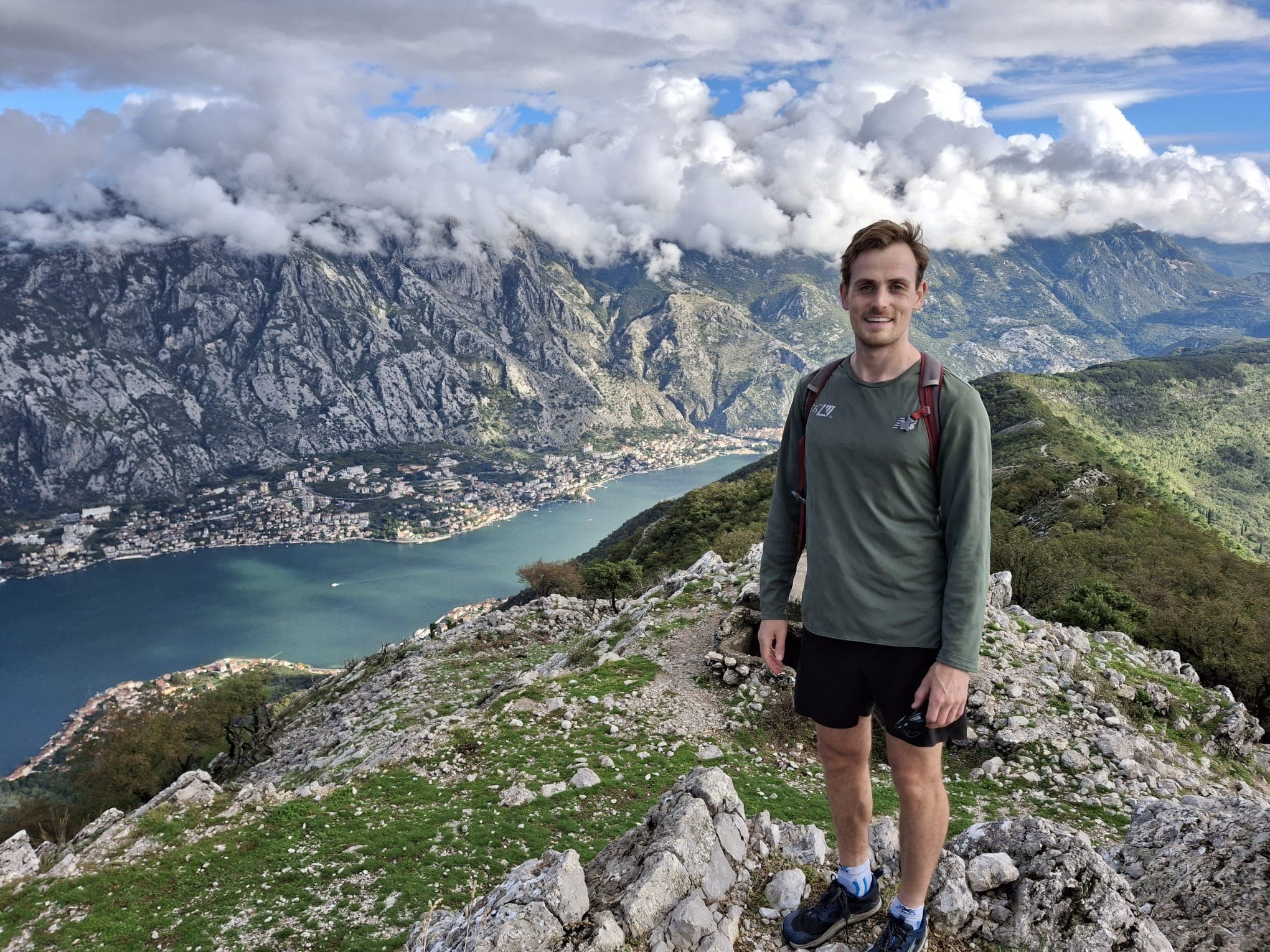 Tom Gray, Physiotherapist at Carter & George, Windsor stands on a rocky mountain trail overlooking a scenic view of a bay, mountains, and clouds.