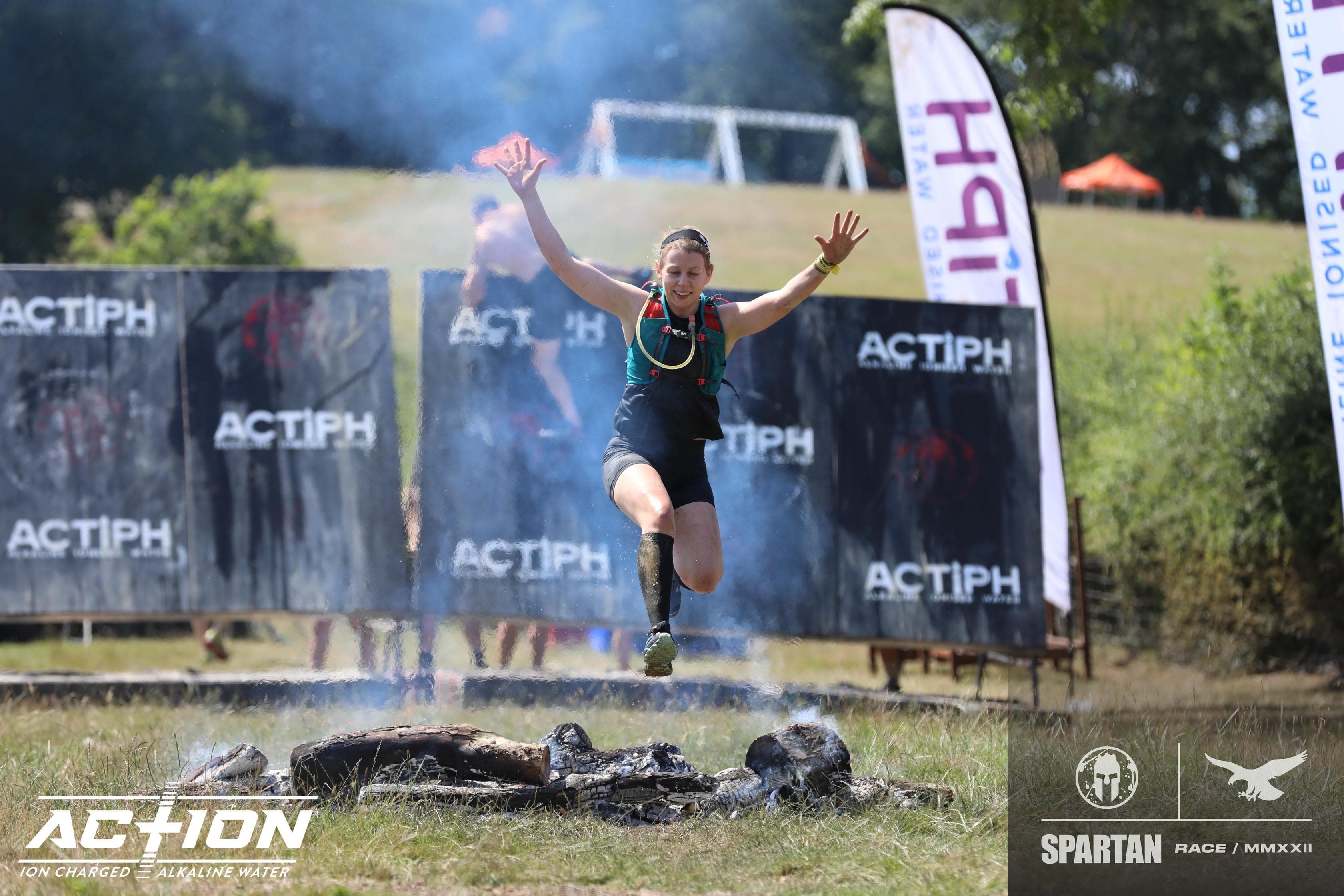 Tara Lawson, Physiotherapist at Carter & George, Wokingham in mid-air jumping over a fire pit during an outdoor obstacle race, with banners and spectators in the background.